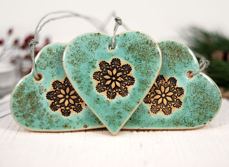May include: Three teal blue ceramic heart ornaments with black snowflake designs. Each heart has a string for hanging.