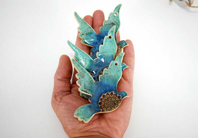 May include: Three ceramic blue and green birds with a mandala design on the bottom. The birds are stacked on top of each other and are being held in a hand.
