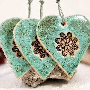May include: Three teal ceramic heart ornaments with black and brown snowflake designs. Each heart has a string loop for hanging.