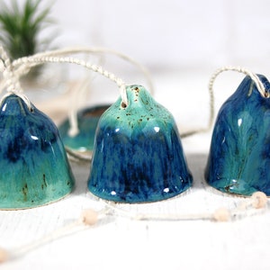 May include: Three ceramic bells in shades of teal and blue, hanging from a white string. The bells are glazed with a mottled finish, giving them a unique and rustic look.