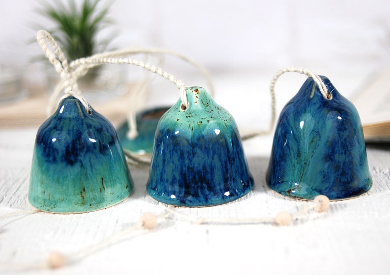 May include: Three ceramic bells in shades of teal and blue, hanging from a white string. The bells are glazed with a mottled finish, giving them a unique and rustic look.