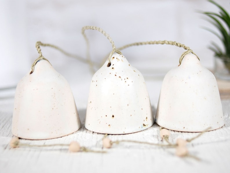 K&ouml;nnte beinhalten: Drei wei&szlig;e Keramikglocken mit gesprenkeltem Finish, die an einer nat&uuml;rlichen Bindfadenkordel h&auml;ngen. Die Glocken sind mit kleinen Holzperlen zusammengebunden.