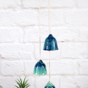May include: A ceramic wind chime with four blue and green bells hanging from a white ceiling. The bells are suspended from a white string with small wooden beads.