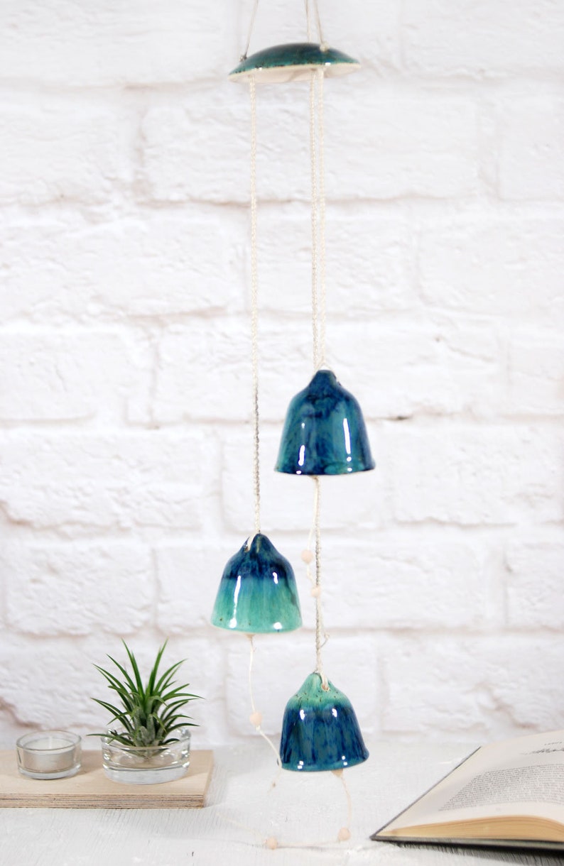 May include: A ceramic wind chime with four blue and green bells hanging from a white ceiling. The bells are suspended from a white string with small wooden beads.
