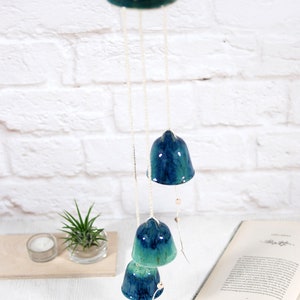 May include: A ceramic wind chime with four blue and green bells hanging from a white string. The bells are decorated with a mottled glaze. The wind chime is hanging from a white ceiling.