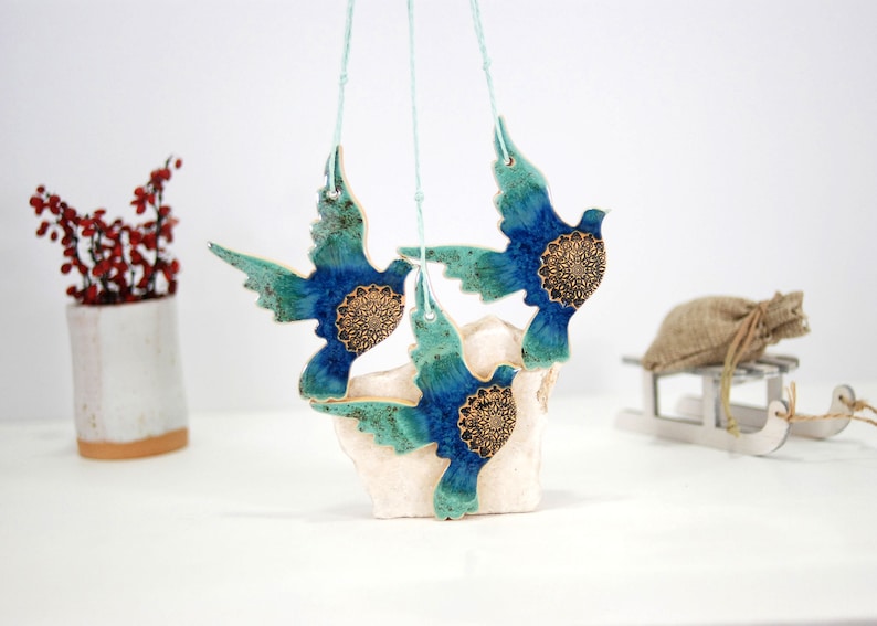 May include: Three blue and green ceramic doves with gold accents hanging from white strings. The doves are suspended from a white surface.