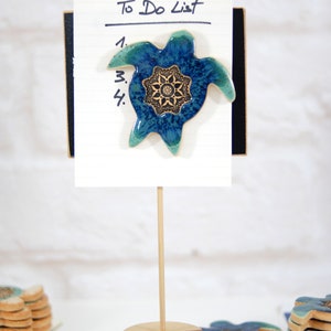 May include: A blue and green ceramic turtle with a gold mandala design is attached to a white to-do list notepad. The notepad is on a wooden stand with a stack of similar turtles on the table.
