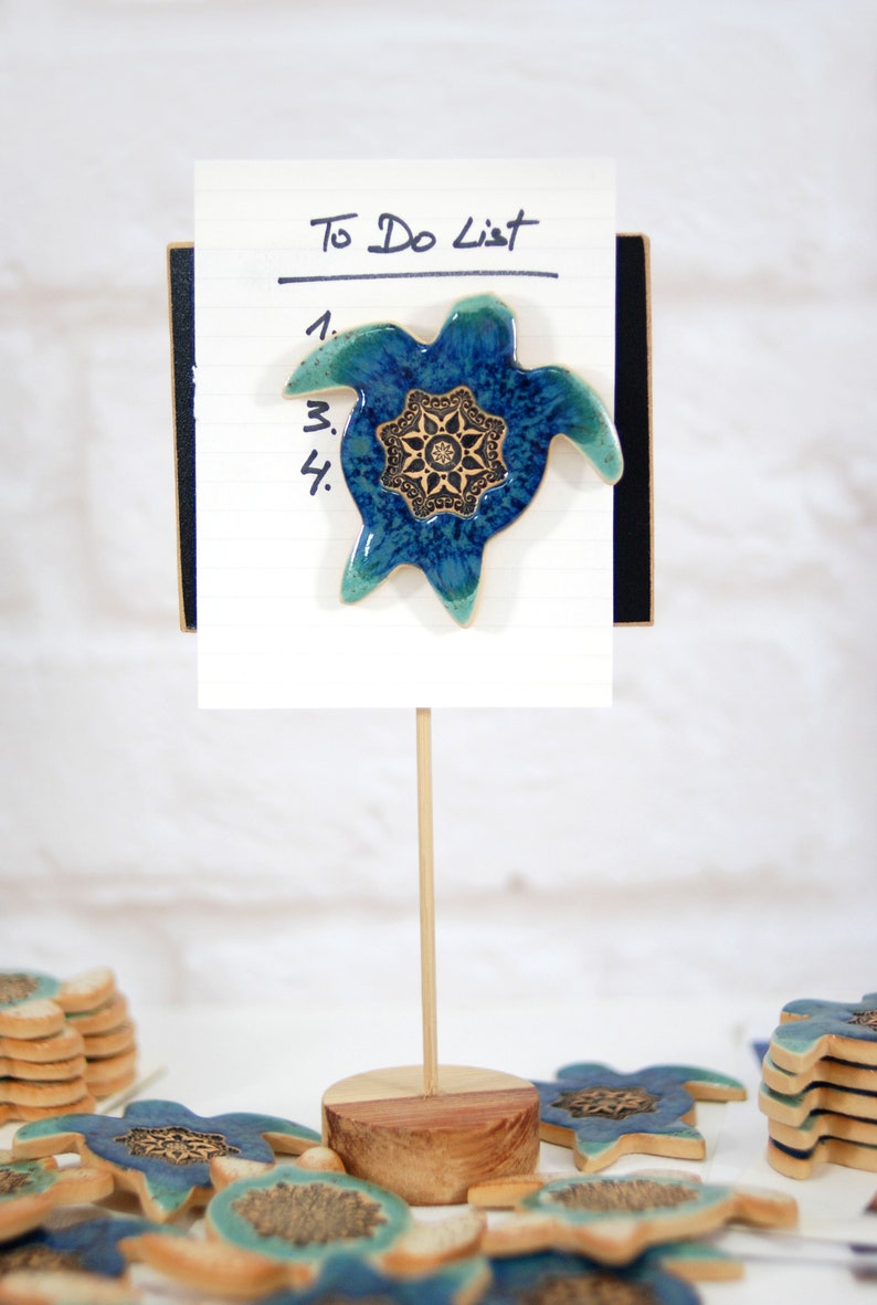 May include: A blue and green ceramic turtle with a gold mandala design is attached to a white to-do list notepad. The notepad is on a wooden stand with a stack of similar turtles on the table.