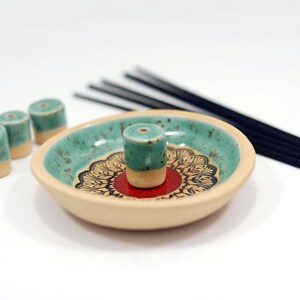 Peut inclure: Un porte-encens en c&eacute;ramique avec un gla&ccedil;age turquoise et cr&egrave;me. Le porte-encens a un motif de mandala rouge et noir et est livr&eacute; avec quatre c&ocirc;nes d'encens turquoise et cr&egrave;me.