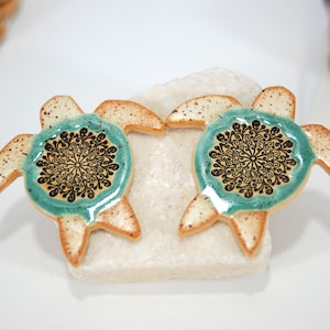 May include: Two ceramic turtle ornaments with a teal glaze and black mandala designs. The turtles are sitting on a white rock.