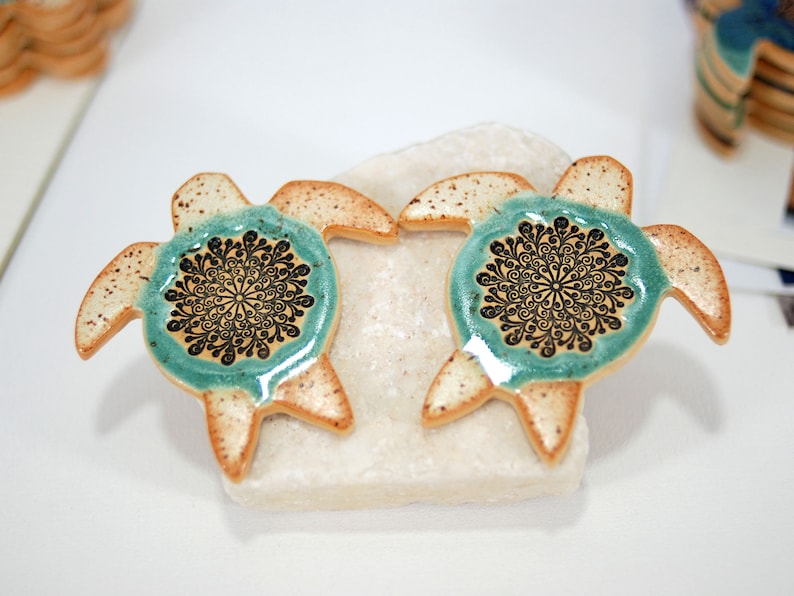 May include: Two ceramic turtle ornaments with a teal glaze and black mandala designs. The turtles are sitting on a white rock.