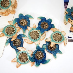 May include: A collection of ceramic turtle ornaments with blue and green glaze and intricate mandala designs. The turtles are arranged in a heart shape on a white surface.