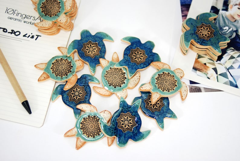 May include: A collection of ceramic turtle ornaments with blue and green glaze and intricate mandala designs. The turtles are arranged in a heart shape on a white surface.