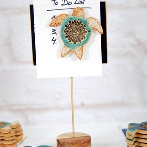 May include: A ceramic turtle with a blue and brown mandala design is attached to a white to-do list notepad with a wooden stand. The notepad has a black backing and the words "To Do List" are written in black ink.