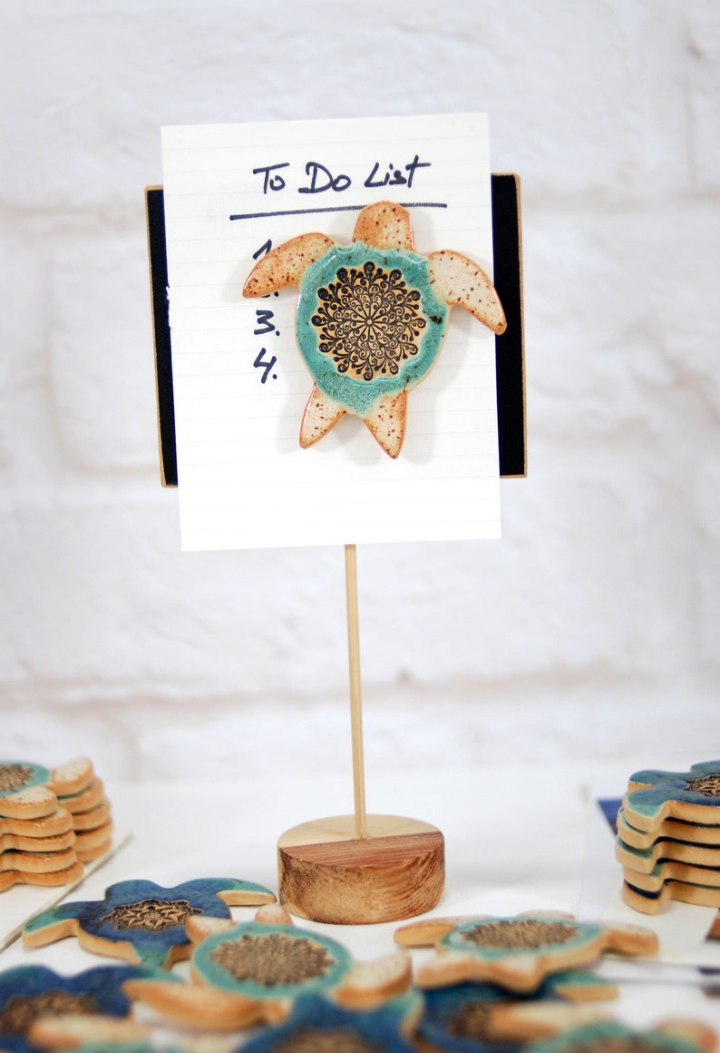 May include: A ceramic turtle with a blue and brown mandala design is attached to a white to-do list notepad with a wooden stand. The notepad has a black backing and the words "To Do List" are written in black ink.