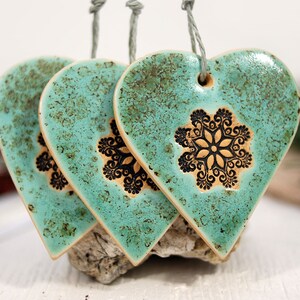 May include: Three teal ceramic heart ornaments with black snowflake designs. The hearts are hanging from twine.