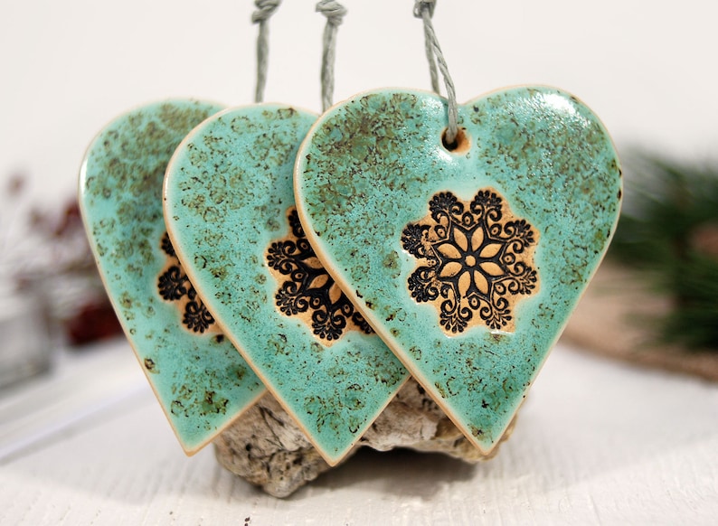 May include: Three teal ceramic heart ornaments with black snowflake designs. The hearts are hanging from twine.