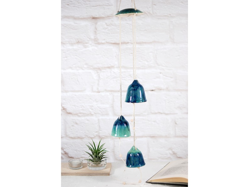 May include: A ceramic wind chime with three blue and green bells hanging from a white string. The bells are suspended from a white ceiling.