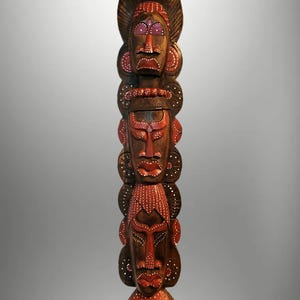 May include: A tall, wooden totem pole featuring three carved faces. The faces are painted with red and white accents on a dark brown background. The top face has a large headdress. The totem is a decorative art piece.