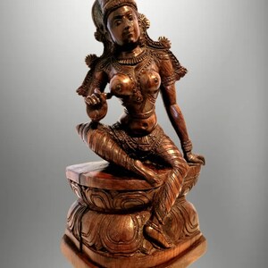 May include: A detailed wooden sculpture of a seated figure with ornate clothing and jewelry. The figure is posed on a carved lotus flower base. The sculpture is a warm brown color and appears to be handcrafted.