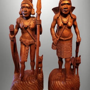 Könnte beinhalten: Zwei Holzskulpturen von Figuren, die jeweils auf einem geschnitzten Sockel stehen. Eine Figur hält einen Speer und einen Stab, mit einem kleinen Hund zu ihren Füßen. Die andere Figur trägt einen Korb und eine Tasche, mit einem Hund zu ihren Füßen.