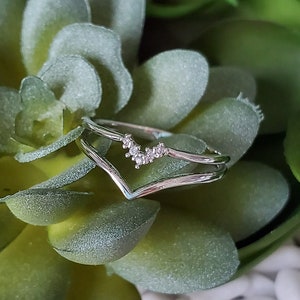 Puede incluir: Un anillo de plata con un diseño en forma de V y pequeñas piedras transparentes engastadas en la banda. El anillo se muestra sobre un lecho de piedras blancas y suculentas verdes.