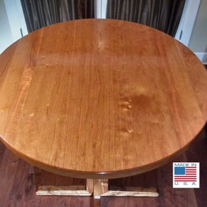 May include: A round wooden table with a natural wood finish. The table has a simple, sturdy base and a smooth, polished top. The table is made in the USA, as indicated by the label on the base.