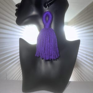 May include: A single, large, purple tassel earring displayed on a black mannequin. The earring is made of thick yarn, with a loop at the top for the hook. The tassel is a deep purple color.