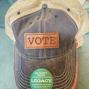 May include: A blue and beige trucker hat with a brown leather patch that says "VOTE" on it. The hat also has a green and white sticker that says "OLD FAVORITE TRUCKER LEGACY" and "PREMIUM, VINTAGE-INSPIRED CAPS FOR A RELAXED LIFESTYLE #LIVEYOURLEGACY".