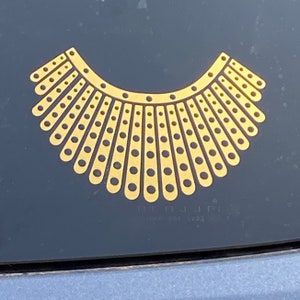 May include: A gold-colored, stylized collar design with a semi-circular shape. The collar features a series of vertical lines with evenly spaced circular cutouts. The design is set against a dark background, possibly a car window.