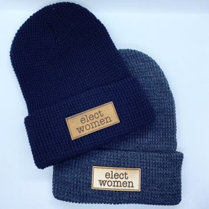 May include: Two knit beanies with the phrase "elect women" on a leather patch. The beanie on top is navy blue and the beanie on the bottom is grey.