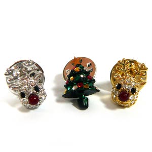 May include: Three Christmas themed brooches. One is a green Christmas tree with red and gold ornaments. The other two are reindeer with red noses and white and gold accents.