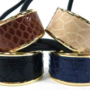 May include: Three hair scrunchies with gold trim and a faux leather finish. The scrunchies are brown, beige, and navy blue.