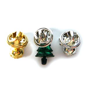 May include: Three Christmas tree pins with different finishes. One is gold, one is silver, and one is green with a silver finish. The pins are all in the shape of a Christmas tree and have a safety pin closure on the back.