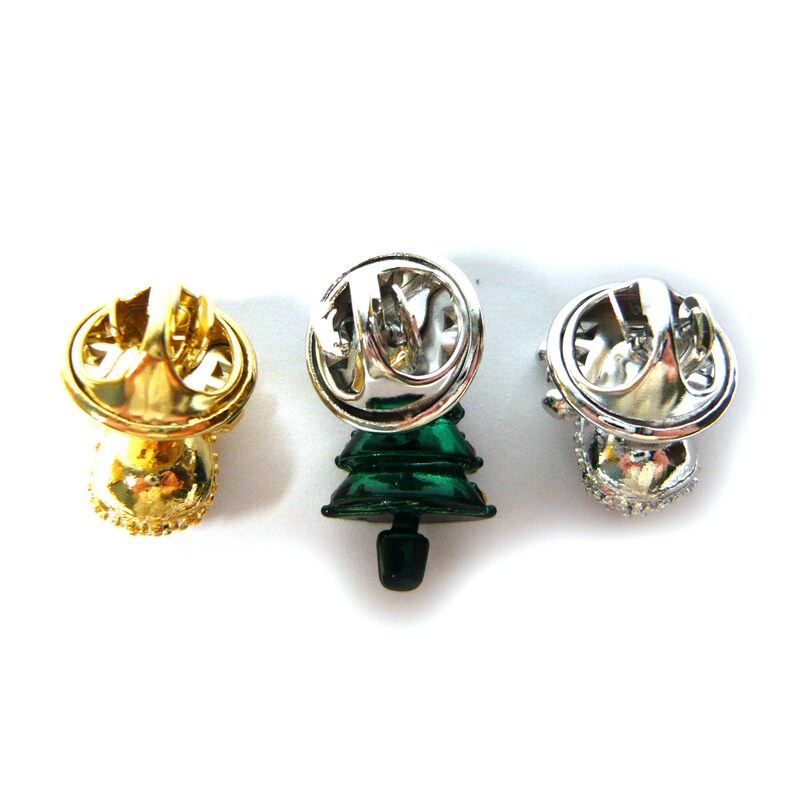 May include: Three Christmas tree pins with different finishes. One is gold, one is silver, and one is green with a silver finish. The pins are all in the shape of a Christmas tree and have a safety pin closure on the back.