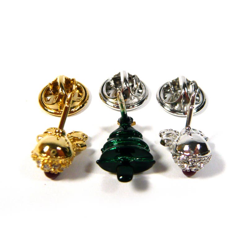 May include: Three Christmas tree pins with different colored finishes. The first pin is gold with clear rhinestones, the second is green with a green enamel finish, and the third is silver with clear rhinestones.