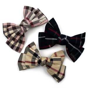 May include: Three plaid fabric hair bows in different colour combinations. The bows are tied in a classic bow shape and have a small tag that says "Hand Made".