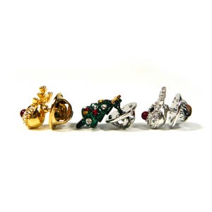 May include: Three gold, silver, and green Christmas themed pins. The pins are shaped like a reindeer, a Christmas tree, and a snowman. Each pin has a red jewel on it.