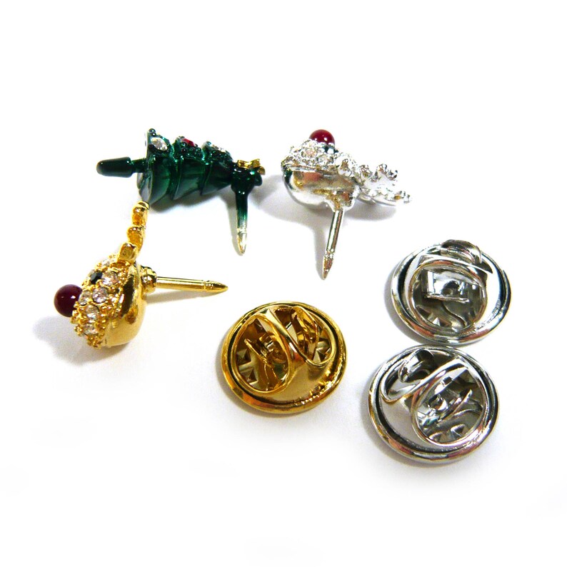 May include: Four Christmas tree pins with red and green enamel and rhinestones. Two pins have gold backs and two have silver backs.