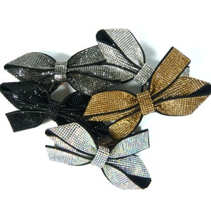 Könnte beinhalten: Vier Haarklammern in Form von Schleifen, jede mit einer anderen Farbe und einem anderen Design. Die Schleifen sind aus schwarzem Samt mit Strasssteinen. Die Farben sind Schwarz, Silber, Gold und schillernd.