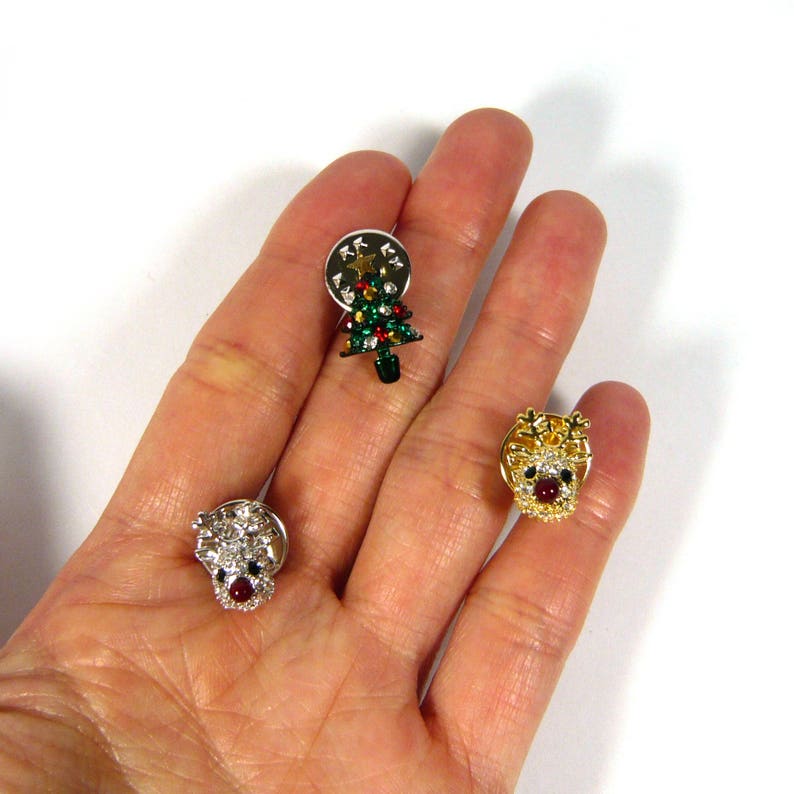 May include: Three Christmas themed brooches. One features a green Christmas tree with red and gold ornaments, another features a gold reindeer with red and white accents, and the last features a white reindeer with red and gold accents.