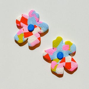 Puede incluir: Un par de pendientes de colores en forma de flor con un centro azul. Los pendientes están hechos de un material texturizado y tienen un esquema de colores rosa, rojo, amarillo y azul.