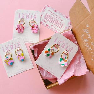 Op de afbeelding: Bloemvormige oorbellen in roze, groen en blauw met gouden ringen, gepresenteerd op kaarten in een bruine doos met roze vloeipapier. De kaarten tonen de tekst "studio IEBIS". De doos heeft de tekst "you are the best!"