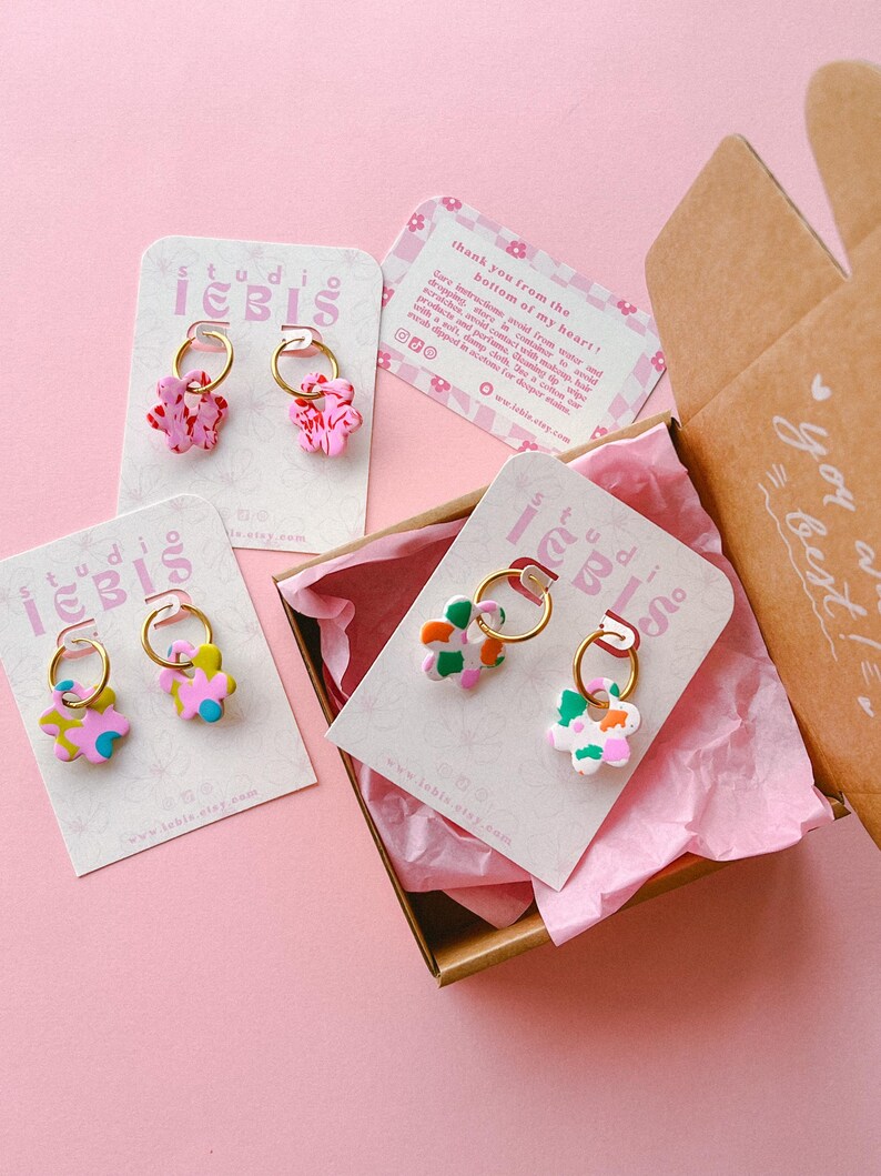 Op de afbeelding: Bloemvormige oorbellen in roze, groen en blauw met gouden ringen, gepresenteerd op kaarten in een bruine doos met roze vloeipapier. De kaarten tonen de tekst "studio IEBIS". De doos heeft de tekst "you are the best!"