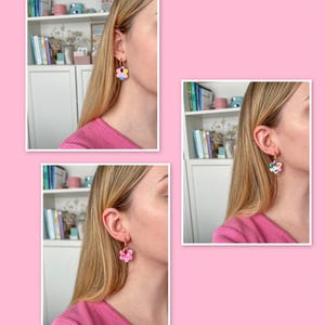 Op de afbeelding: Gouden oorringen met bloemvormige hangers in roze, geel, blauw en groen. De oorbellen worden getoond aan een oor, tegen een roze achtergrond.
