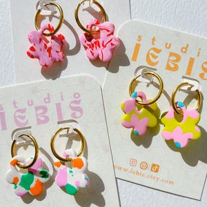 Op de afbeelding: Handgemaakte bloemvormige oorbellen in verschillende kleuren, waaronder roze, wit, groen, oranje en geel. Elke oorbel heeft een gouden ring. De oorbellen worden gepresenteerd op kaarten met de tekst "studio iebis" en een websiteadres.