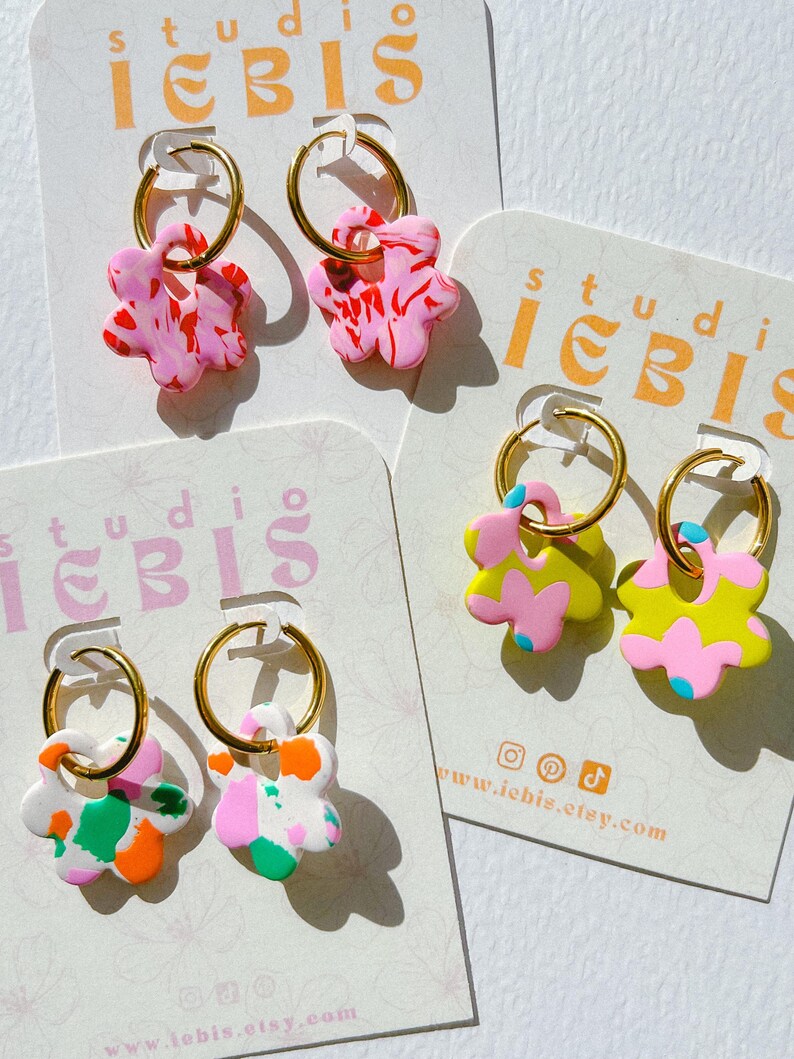 Op de afbeelding: Handgemaakte bloemvormige oorbellen in verschillende kleuren, waaronder roze, wit, groen, oranje en geel. Elke oorbel heeft een gouden ring. De oorbellen worden gepresenteerd op kaarten met de tekst "studio iebis" en een websiteadres.