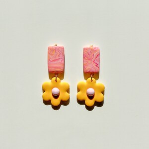 May include: A pair of dangle earrings with a pink and yellow marbled rectangle charm and a yellow flower charm with a pink center.