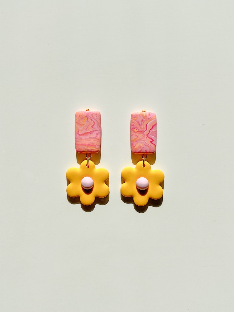May include: A pair of dangle earrings with a pink and yellow marbled rectangle charm and a yellow flower charm with a pink center.