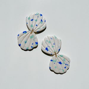 Op de afbeelding: Een paar witte oorbellen in de vorm van een zeeschelp met blauwe en groene spikkels. De oorbellen zijn verbonden door een kleine goudkleurige lus. De oorbellen hebben een getextureerd oppervlak en een geschulpte rand.